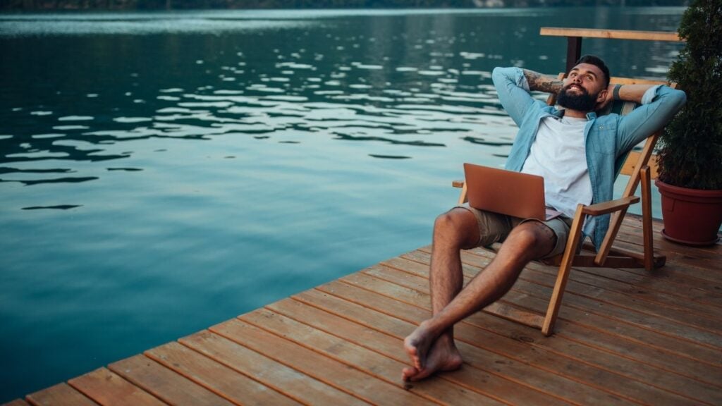 Homme assis sur une chaise longue au bord d'un lac en train de se détendre. Un ordinateur portable est posé sur ses genoux.
