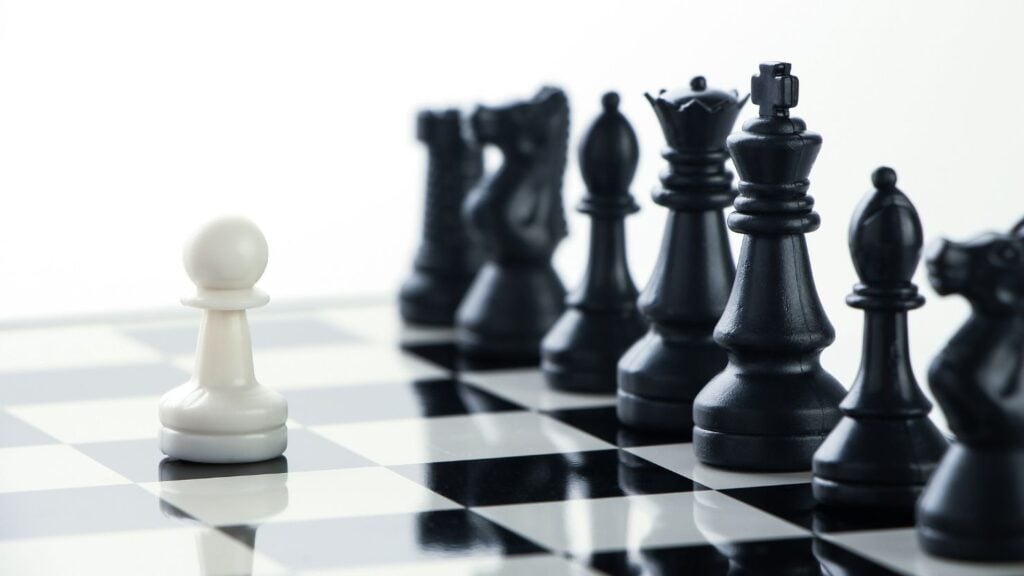 Chess pieces on a board with a white pawn facing a row of black chess pieces