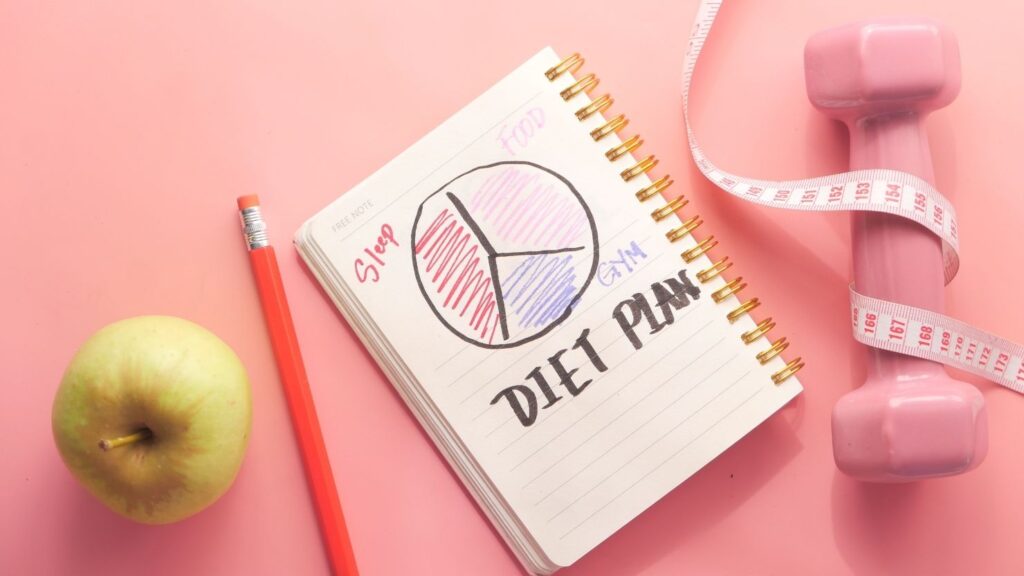 An image of an apple, a notebook and pencil, and a dumbbell with a tape measure wrapped around it to represent a "diet plan".