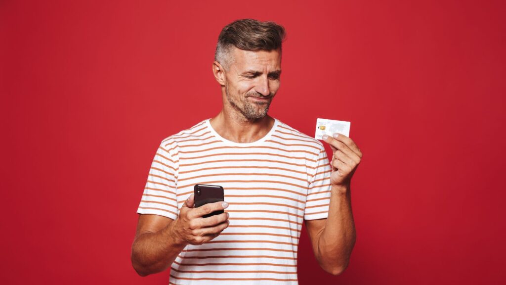 a portrait of a disappointed man holding his phone while looking at his credit card