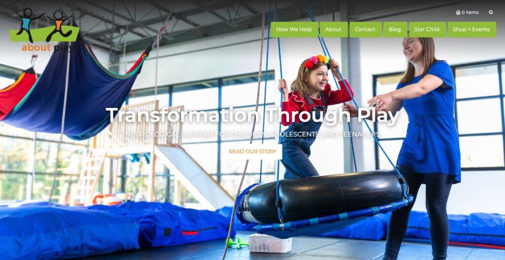 Girl in a red sweater enjoys a suspended swing ride with an adult helper in a bright indoor playground, conveying playful therapy and support through play.