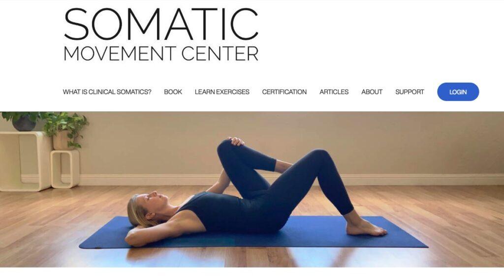 Woman in a black athletic outfit lying on a blue yoga mat while lifting her hips in a bridge pose in a bright studio.